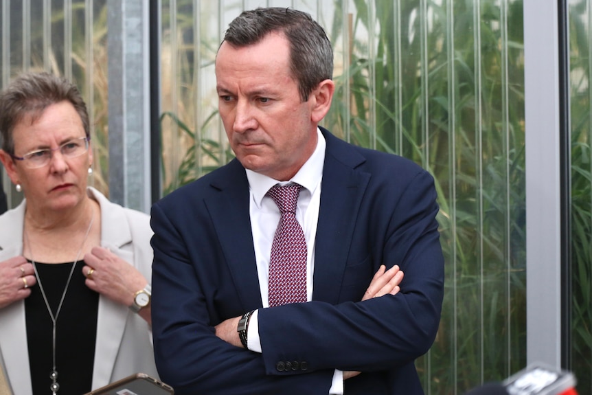 A mid-shot of WA Premier Mark McGowan looking downwards with his arms folded as he listens during a media conference.