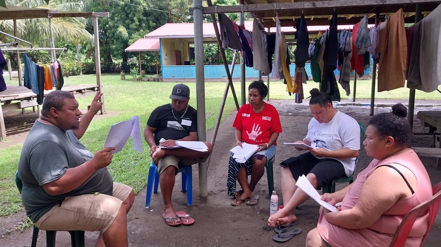 Group of actors sit outdoors on plastic chairs in PNG village reading script. 