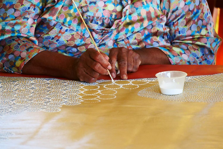 A close-up of an Aboriginal woman's hands as she paints a dot painting of a yellowbelly fish.