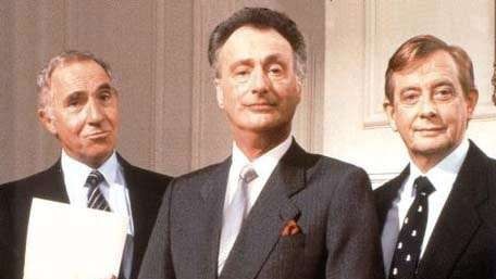 The three main characters from British political satire Yes Minister stand together wearing suits.