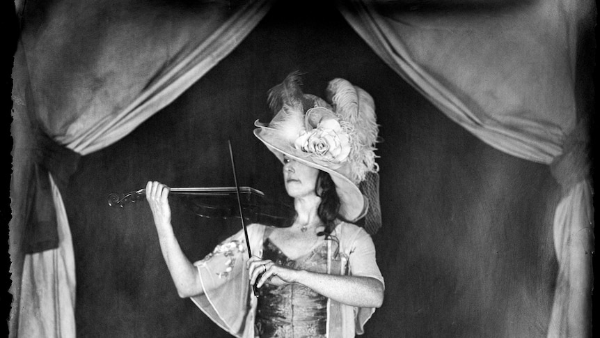 Black and white wet plate (historical looking) photo of Shane wearing a big ornate hat and playing a violin. 