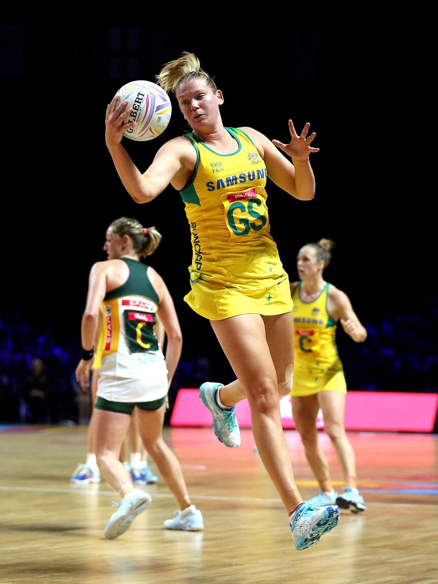 A very tall woman in an Australian netball jersey handles the ball while leaping through the air