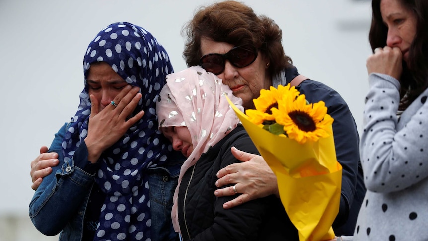 A woman wearing a headscarf wipes away a tear with a hand as she embraces another woman.