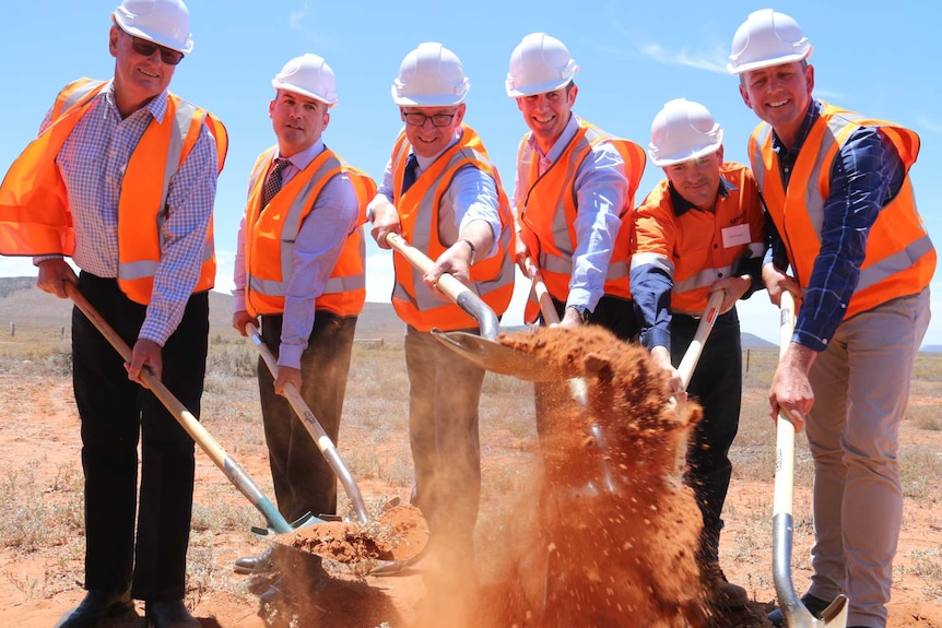 Six men in fluorescent jackets and hard hats hold shovels.