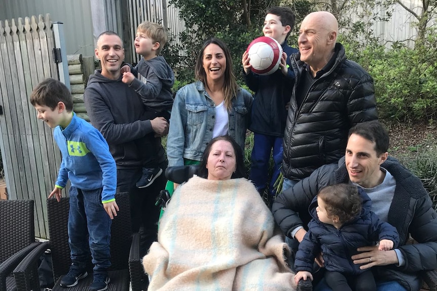 Woman sits in wheelchair with blanket tucked up around her neck surrounded by her smiling husband, children and grandchildren