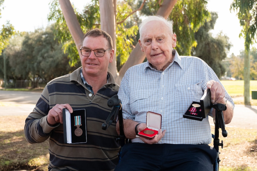 old man sitting on walker and man with glasses kneeling next to him both holding medals