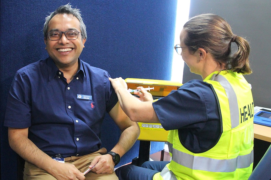 Dr Gulam Khandaker sitting in a chair smiling while a worker injects the COVID-19 vaccine.