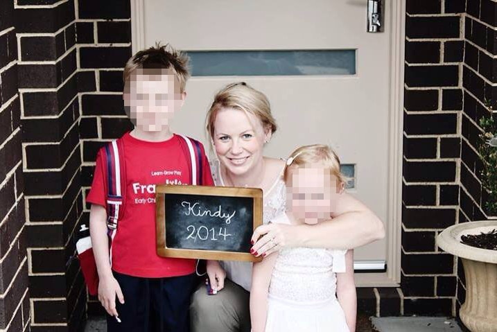 A woman sitting in front of a door with a young child on either side. Holding a sign that says Kindy 2014.