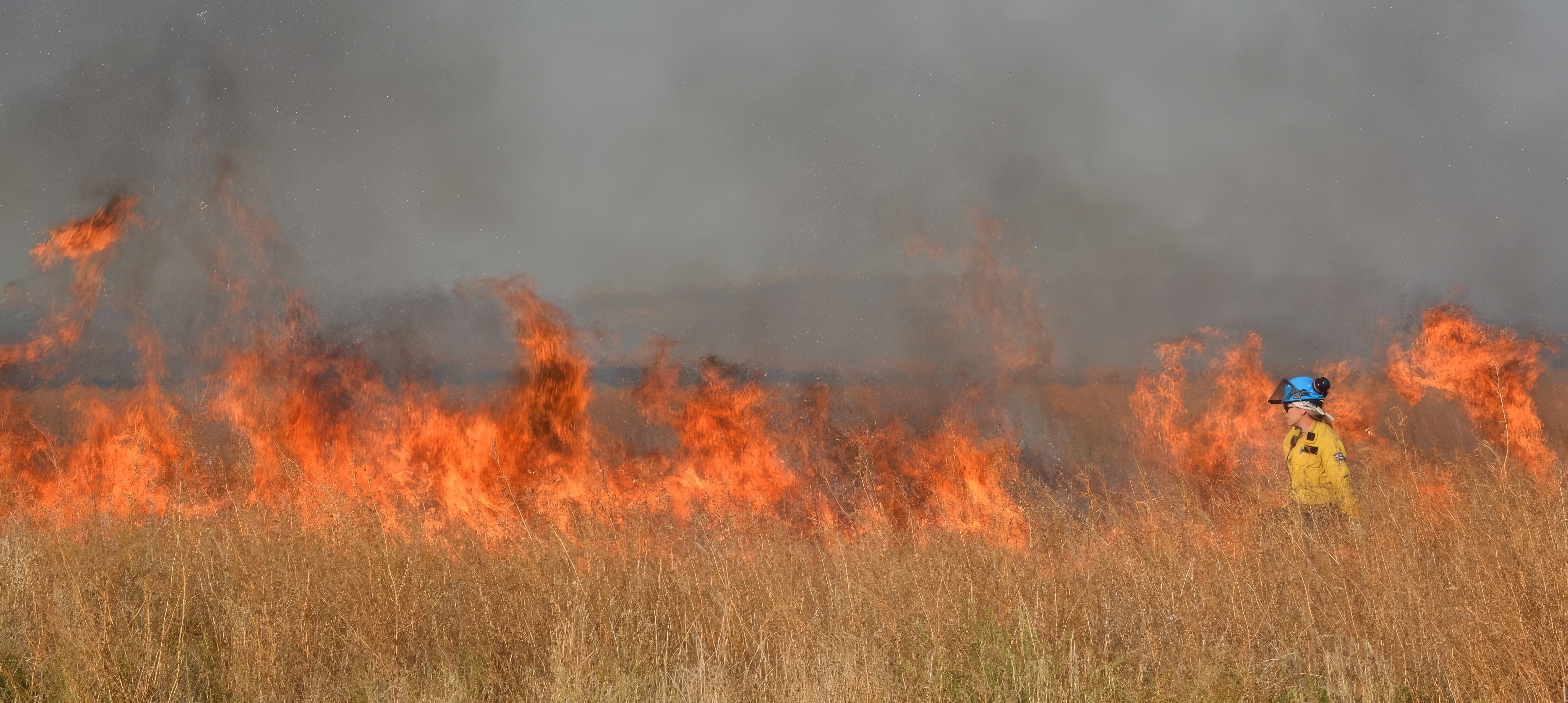 Uma mulher vestida com uniforme de bombeiro caminha em frente às chamas. 