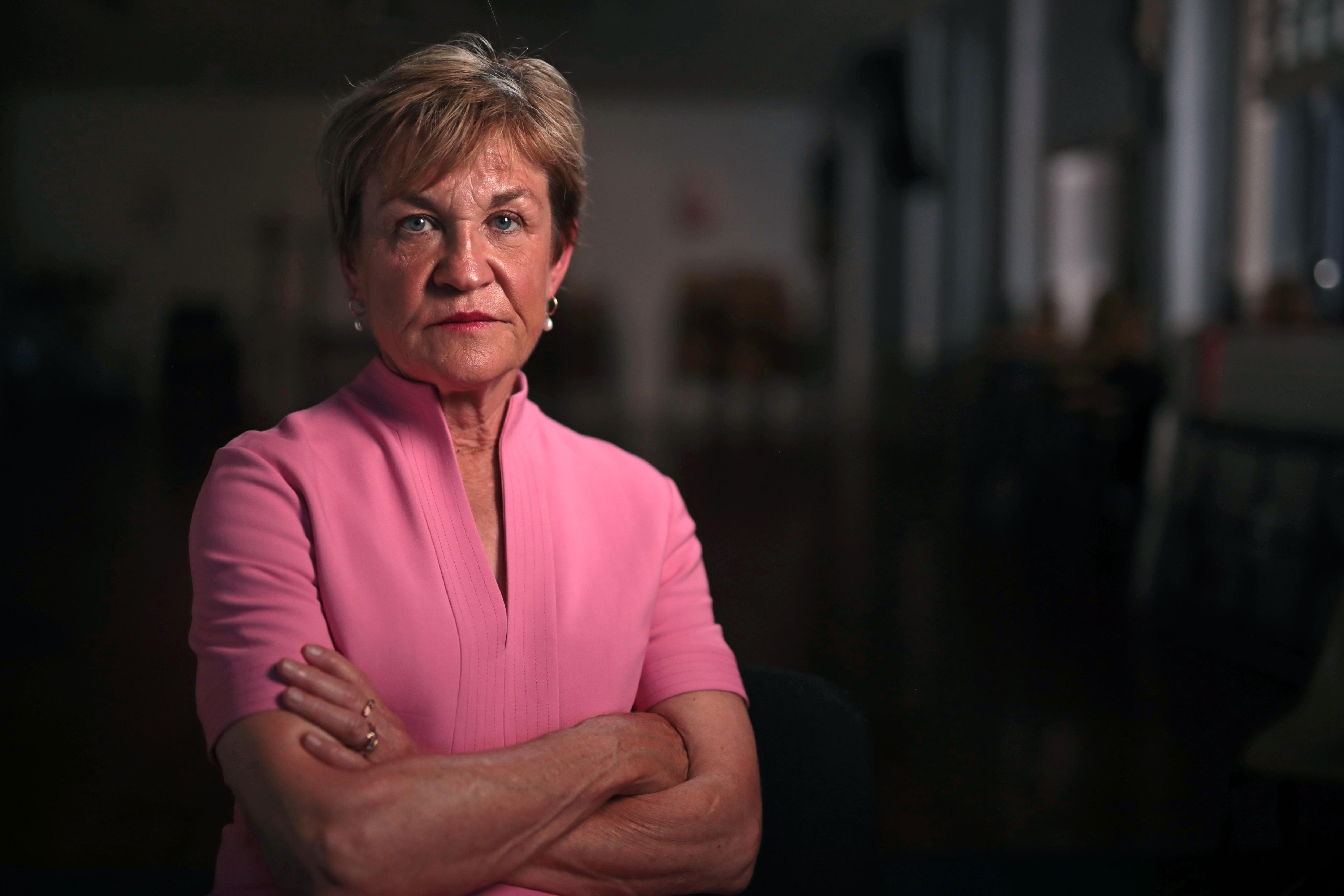 A woman, arms folded, stares at the camera with a determined expression. She is wearing pink, the room behind is dark.