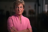 A woman, arms folded, stares at the camera with a determined expression. She is wearing pink, the room behind is dark.