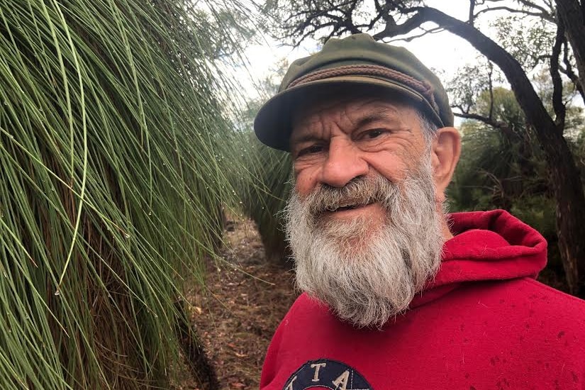 Len Collard wears a red jumper and smiles at the camera, standing next to a grass tree in bushland.