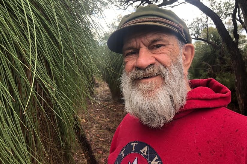 Len Collard wears a red jumper and smiles at the camera, standing next to a grass tree in bushland.