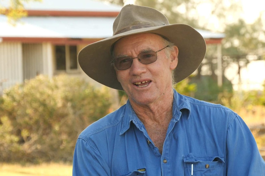 Brendan McNamara, a western Queensland grazier, in focus wearing a hat and blue shirt, with house our of focus behind him