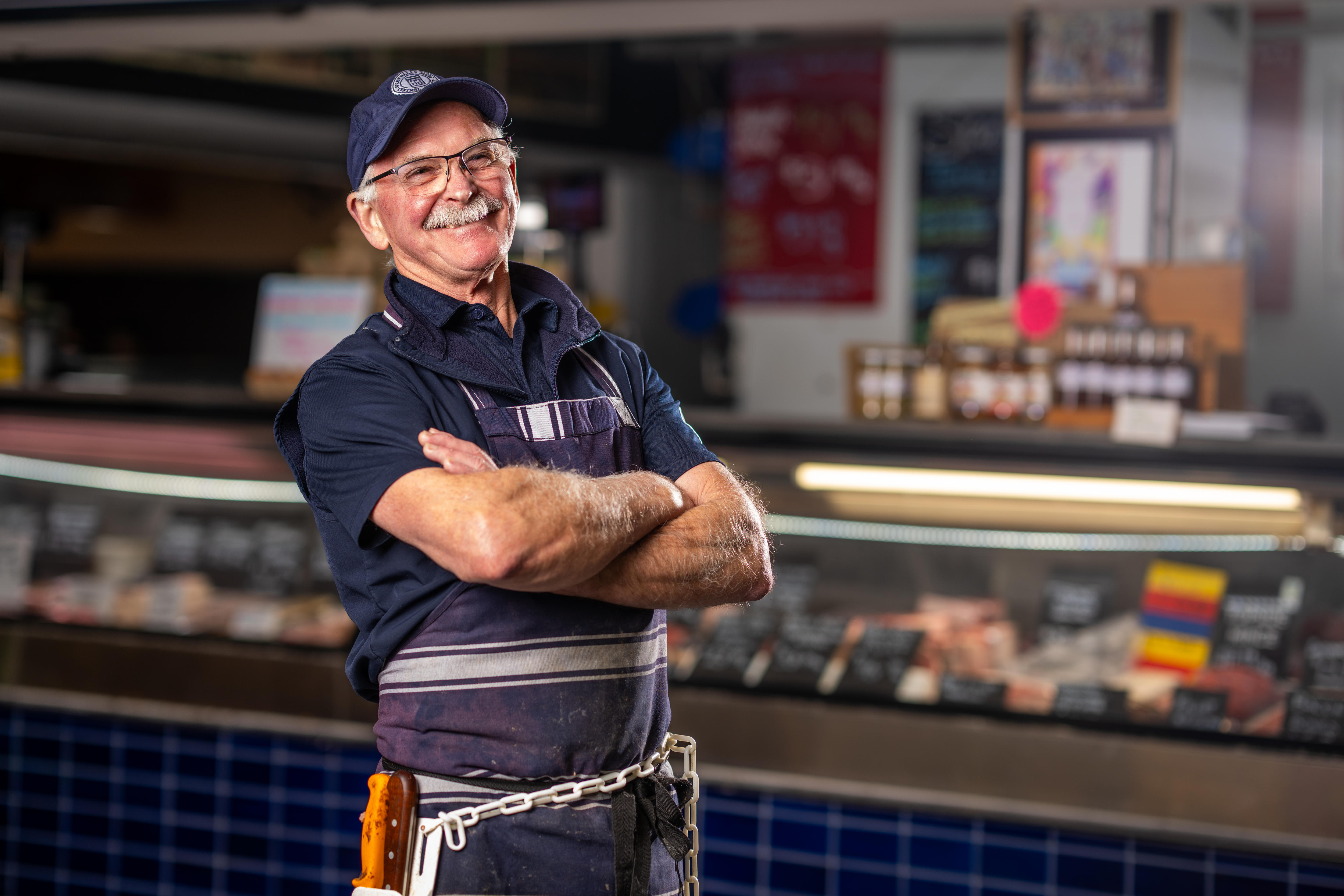 A man with grey moustache and in navy blue with a cap, shirt and apron in front of butcher counter. 