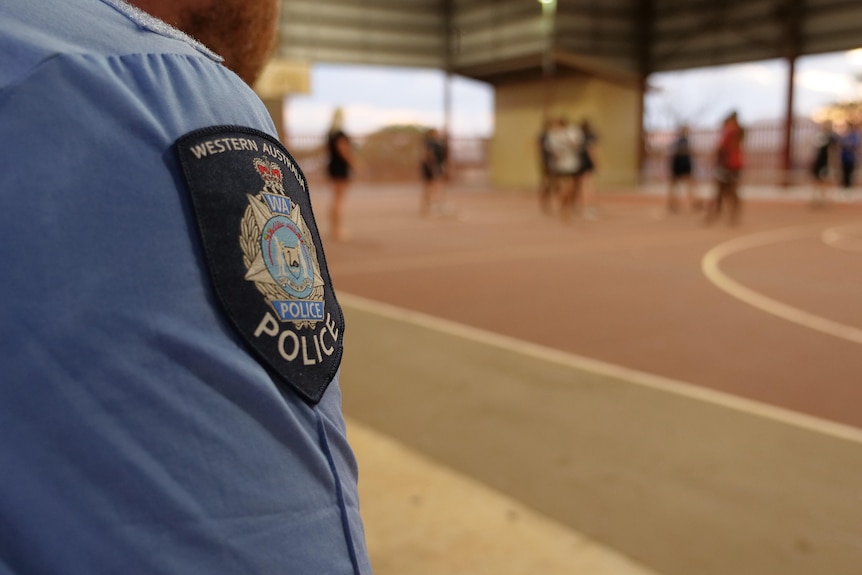 A police officer's badge on his arm is visible as he watches children playing basketball