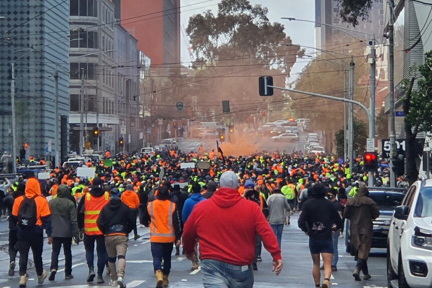 A crowd of hundreds, many in hi-vis jackets, march through Melbourne's CBD with an orange flare haze in the air.