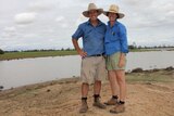 A couple in farm work clothes stand on a hill smiling with a big lake of water and green grass behind them