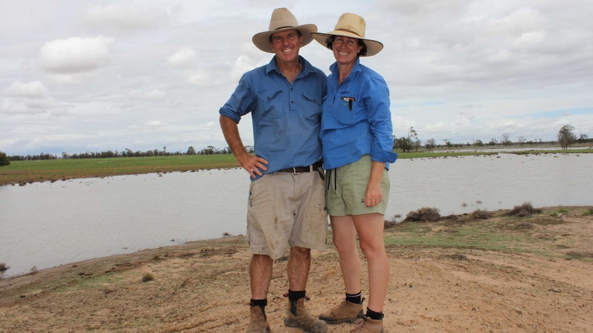 A couple in farm work clothes stand on a hill smiling with a big lake of water and green grass behind them