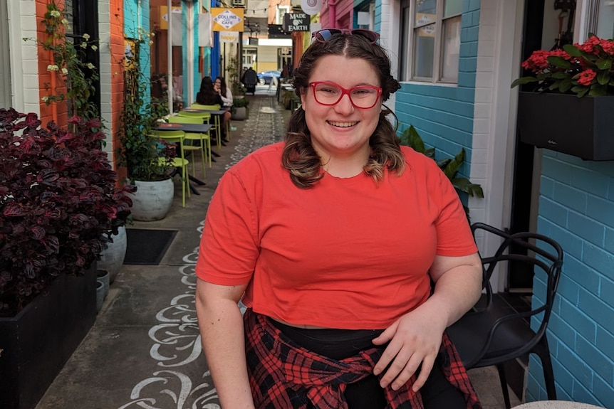 A woman with pink glasses smiles as she stands in a cafe alleyway. 