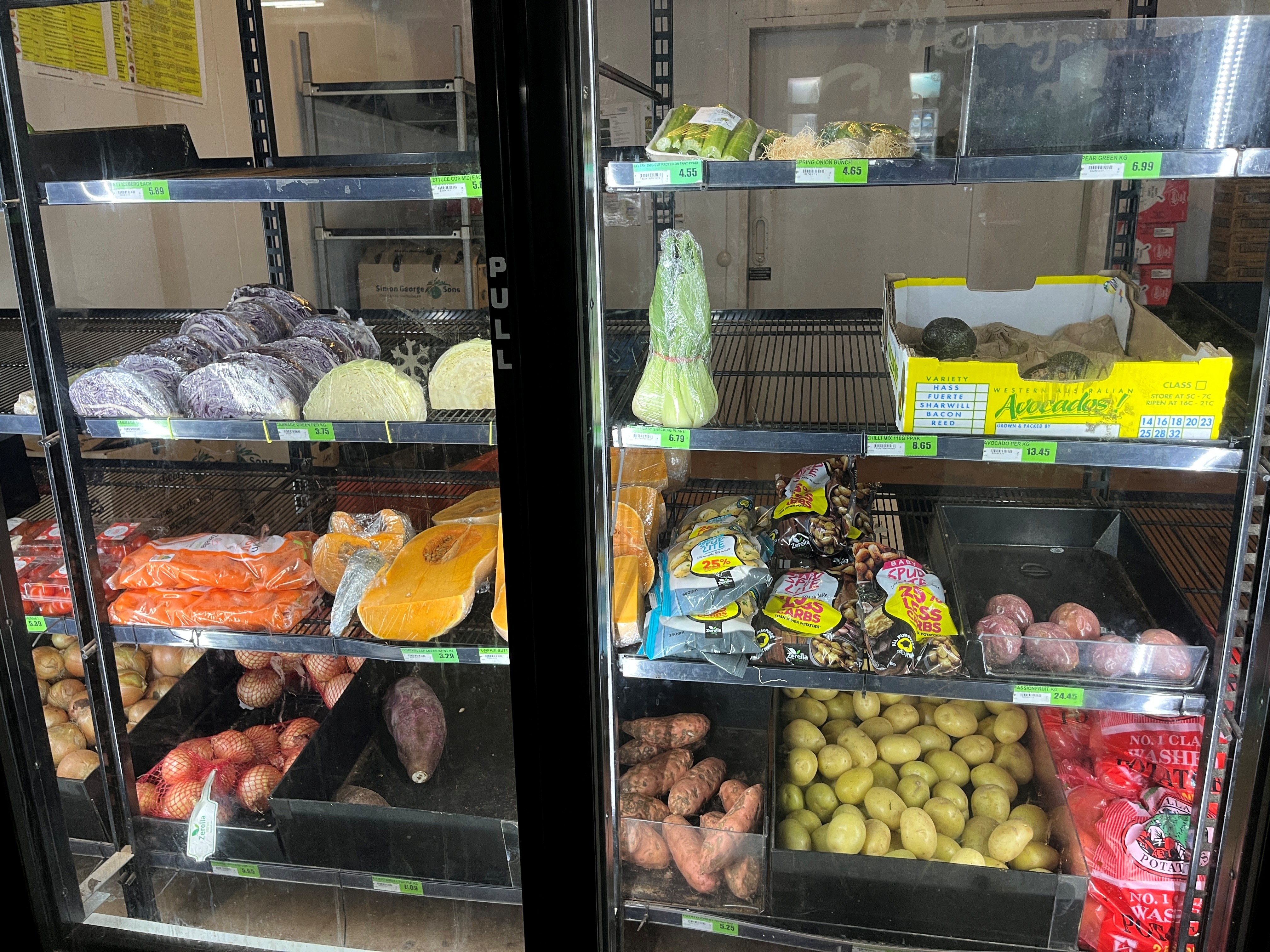 A photo shows fresh produce, including potatoes and pumpkins, behind a glass fridge door in a small supermarket.