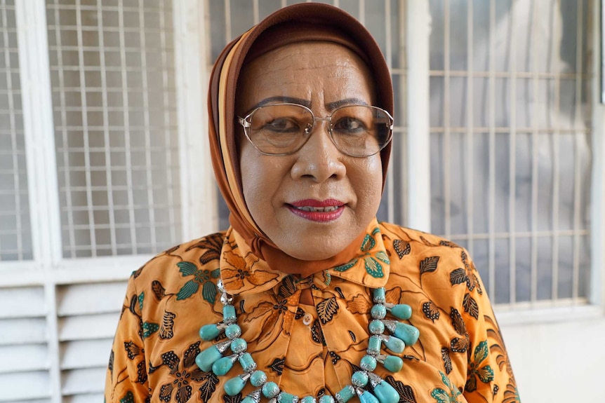 A transgender woman wearing a yellow floral hijab and a blue necklace, bright lipstick.