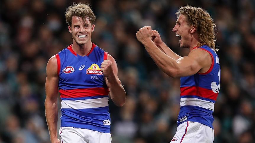 A grinning AFL player pumps his fist, while a teammate shouts at him in joy after a goal.