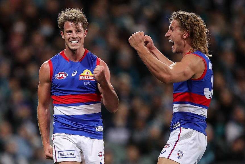 A grinning AFL player pumps his fist, while a teammate shouts at him in joy after a goal.