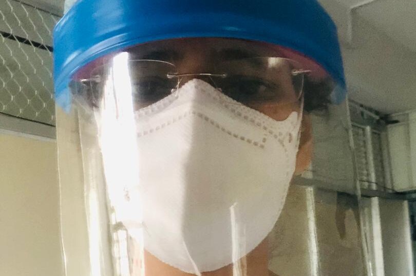 A woman wearing a hair net, face shield and mask and glasses takes a selfie.