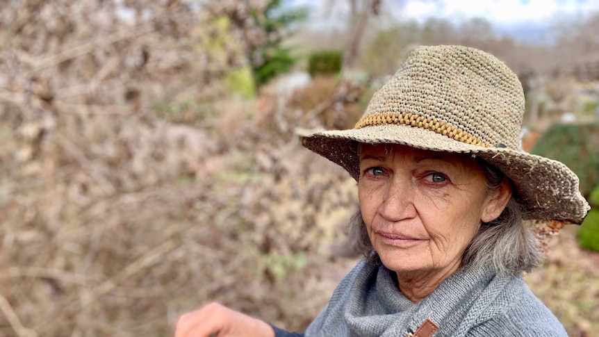 Middle aged lady wearing a straw hat and grey top with brown, dead plants in her garden.
