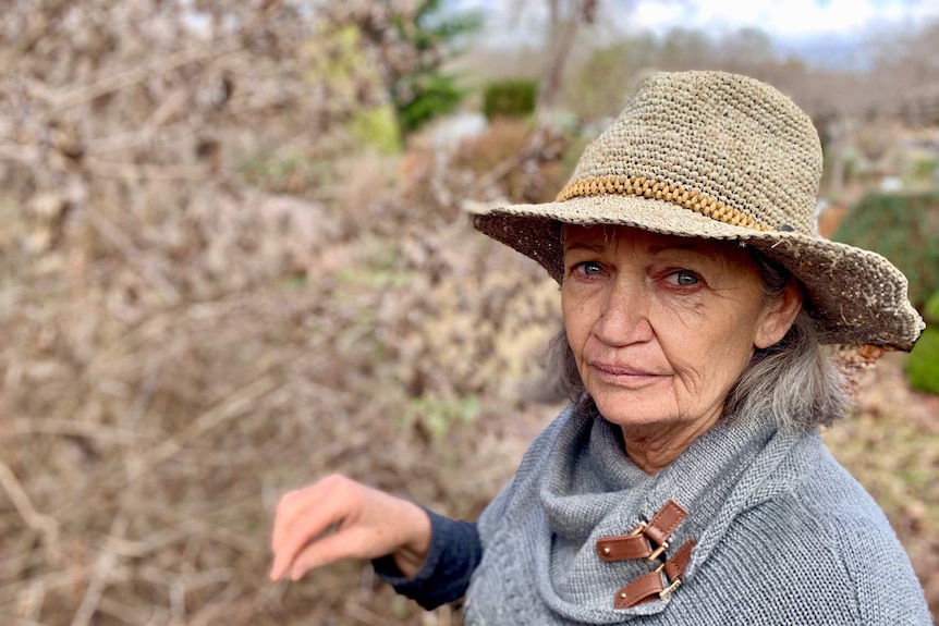 Middle aged lady wearing a straw hat and grey top with brown, dead plants in her garden.