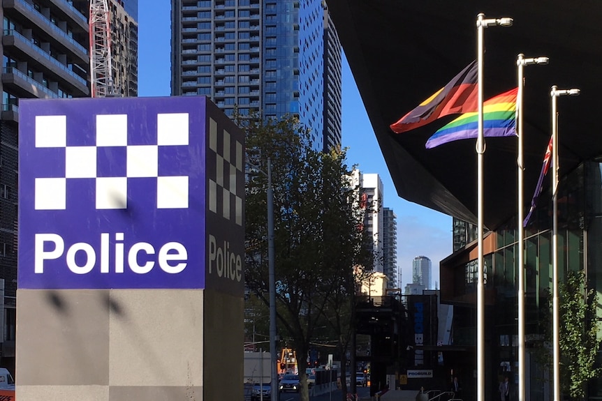 A  rainbow flag flies between the Aboriginal and Australian flags, behind a large 'police' sign.