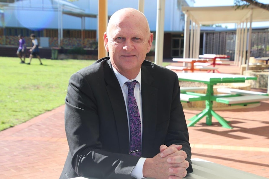 Wayne sits at a bench in a school yard in the sunshine, wearing a suit with his hands clasped in front of him.