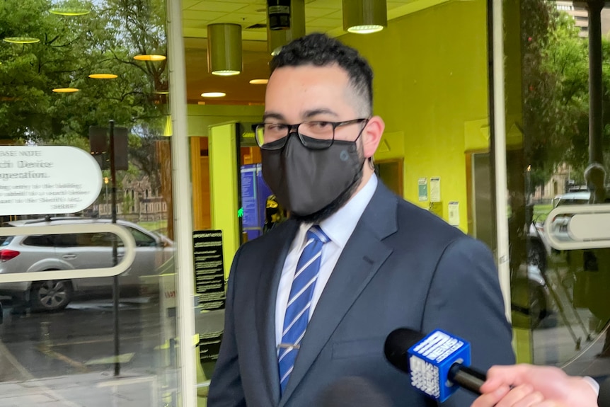 A man wearing a navy suit, white shirt, blue tie and a black face mask walks outside court. Microphones are pointed towards him