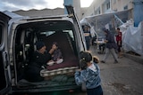A family seeks shelter in the back of a van in the street as a father feeds his baby a spoonful of food.