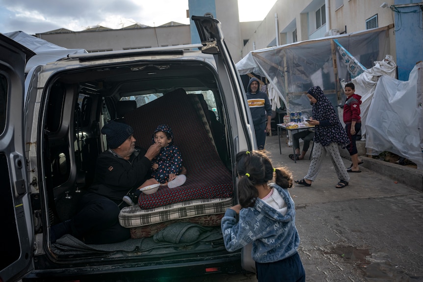 A family seeks shelter in the back of a van in the street as a father feeds his baby a spoonful of food.
