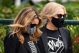 Dannii Minogue and Kylie Minogue, wearing masks and dressed in black with Mushroom-branded T-shirts, walk together.