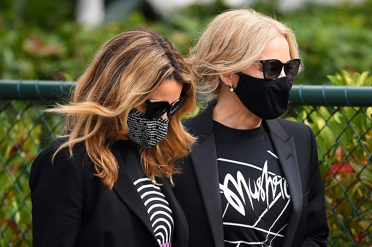 Dannii Minogue and Kylie Minogue, wearing masks and dressed in black with Mushroom-branded T-shirts, walk together.