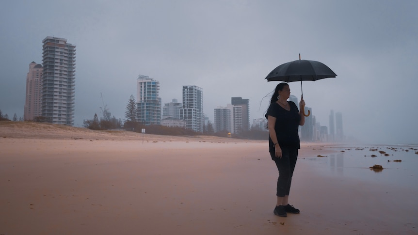 A woman holds an umbrella and stands on a beach in the rain. A cityscape is visible behind her.