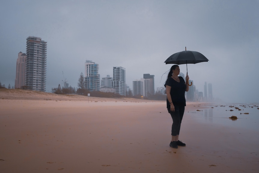 A woman holds an umbrella and stands on a beach in the rain. A cityscape is visible behind her.
