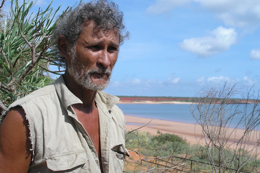 A man with curly grey hair and a beard stands in front of a remote beach.
