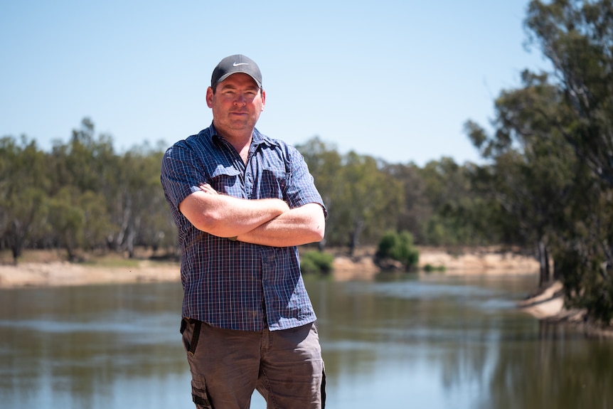 A man is a short-sleeved blue shirt stands with arms crossed in front of the Murray River.