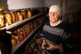 An older woman with a cream scarf wrapped around her head stands next to jars of picked food