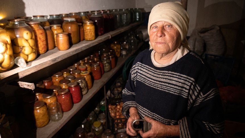 An older woman with a cream scarf wrapped around her head stands next to jars of picked food