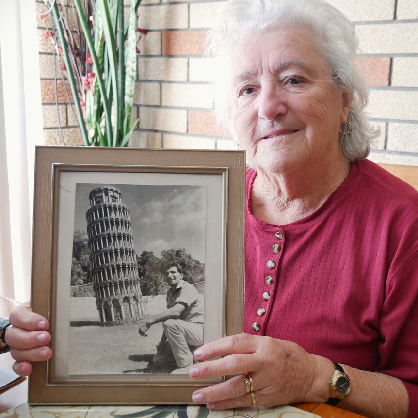 A women holds a framed photo of a man kneeling near a leaning tower. 