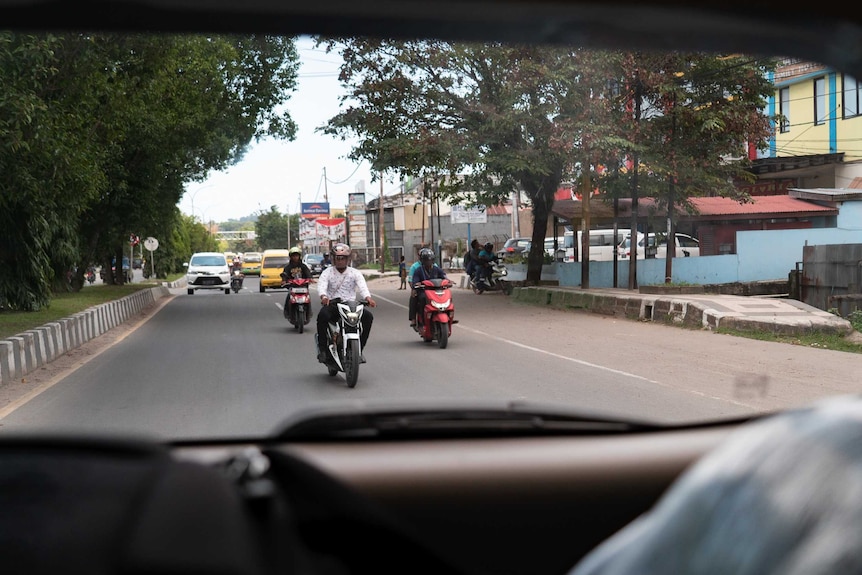 Through a car's rear window, men on scooters are seen driving on a street.