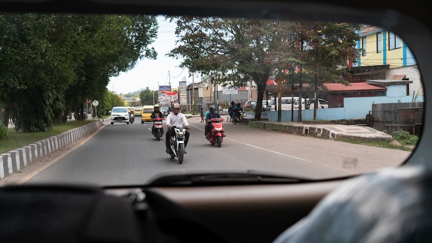 Through a car's rear window, men on scooters are seen driving on a street.