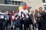 A man in a light pink shirt speaks into microphones with an Aboriginal flag and crowd of supporters behind him