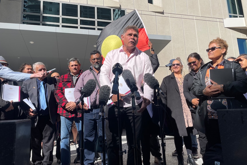 A man in a light pink shirt speaks into microphones with an Aboriginal flag and crowd of supporters behind him