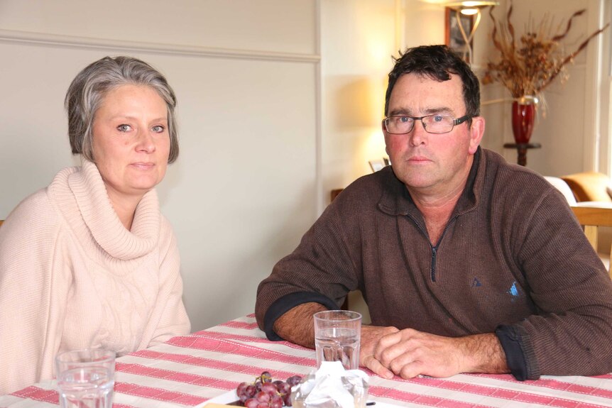A late middle-aged couple sit at a dining table with sombre expressions on their faces.